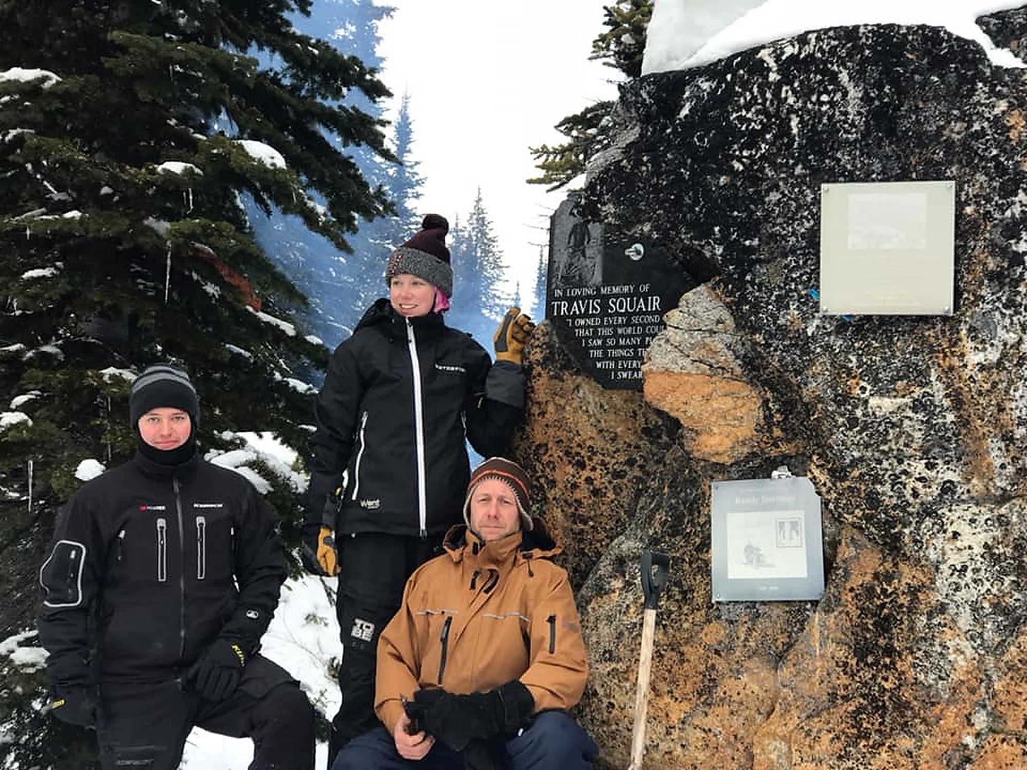 Three people standing at a granite memorial set into a mountain rock face in the snow